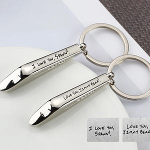Puede incluir: Dos llaveros plateados con una etiqueta rectangular y un extremo redondeado. Las etiquetas están grabadas con las frases "I LOVE YOU, SPAWN!" y "LOVE YOU, JIMMY BEAR!". Cada llavero tiene un anillo plateado.