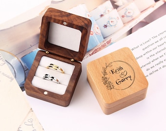 May include: A wooden ring box with two rings inside. The box is engraved with the names "Keith & Garry" and a floral design. The box is made of light brown wood.