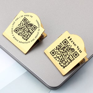 Könnte beinhalten: Zwei goldfarbene Metall-Binderclips mit einem schwarzen QR-Code und Text. Der obere Clip sagt "Ich liebe dich". Der untere Clip sagt "Sobald ich dich sah, wusste ich, dass ein großes Abenteuer bevorstand".