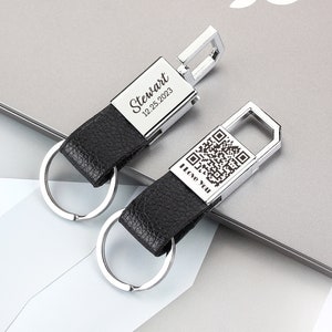 Op de afbeelding: Twee zwarte leren sleutelhangers met zilveren metalen sleutelringen. De ene sleutelhanger heeft de naam "Stewart" en de datum "12.25.2023" erop gegraveerd. De andere sleutelhanger heeft een QR-code en de woorden "I Love You" erop gegraveerd.