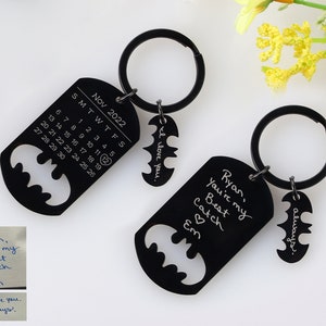 May include: Two black metal keychains with a Batman silhouette design. One keychain features a calendar with the month of November 2022 and the text "I love you." The other keychain has the text "Ryan, you're my best catch Em" and "always".