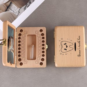May include: A wooden tooth box with a hinged lid, open to reveal a compartment for baby teeth and a small vial. The lid features a cartoon tooth design and the text "Bruce's tooth box". The box is made of light-colored wood.