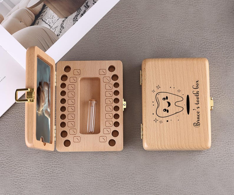 May include: A wooden tooth box with a hinged lid, open to reveal a compartment for baby teeth and a small vial. The lid features a cartoon tooth design and the text "Bruce's tooth box". The box is made of light-colored wood.
