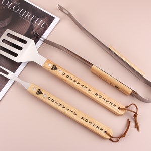 Puede incluir: Un juego de tres herramientas de barbacoa de plata con mangos de madera. Las herramientas incluyen una espátula, un tenedor y unas pinzas. Los mangos están grabados con el texto "BBQ ODVALA ROTSSEN".