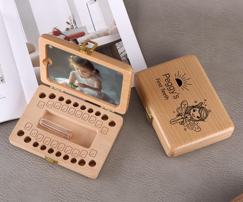 May include: Wooden baby tooth keepsake box with a hinged lid. The lid has a photo of a child. The box is open, revealing spaces for baby teeth and a small vial. The closed box is engraved with "Peggy's First Teeth" and a fairy illustration.