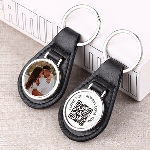 Puede incluir: Dos llaveros de cuero negro con anillos de metal plateado. Un llavero tiene un inserto de foto redondo con una pareja. El otro llavero tiene un inserto redondo con un código QR y el texto "I LOVE YOU. I ALWAYS LOVE YOU."
