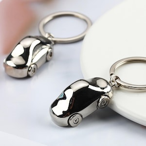 K&ouml;nnte beinhalten: Zwei silberne Autoschl&uuml;sselanh&auml;nger. Jeder Schl&uuml;sselanh&auml;nger hat ein Miniatur-Autodesign mit R&auml;dern und einem Schl&uuml;sselring. Die Schl&uuml;sselanh&auml;nger sind reflektierend, mit einer glatten, polierten Oberfl&auml;che. Der Hintergrund ist ein weiches Wei&szlig; und Rosa.
