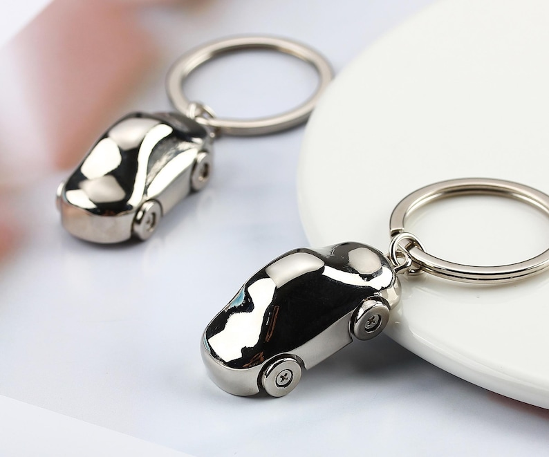K&ouml;nnte beinhalten: Zwei silberne Autoschl&uuml;sselanh&auml;nger. Jeder Schl&uuml;sselanh&auml;nger hat ein Miniatur-Autodesign mit R&auml;dern und einem Schl&uuml;sselring. Die Schl&uuml;sselanh&auml;nger sind reflektierend, mit einer glatten, polierten Oberfl&auml;che. Der Hintergrund ist ein weiches Wei&szlig; und Rosa.
