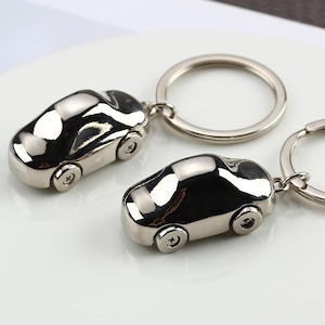 K&ouml;nnte beinhalten: Zwei silberne Autoschl&uuml;sselanh&auml;nger. Jeder Schl&uuml;sselanh&auml;nger zeigt ein Miniaturauto mit detaillierten R&auml;dern und einer glatten, reflektierenden Oberfl&auml;che. Die Schl&uuml;sselanh&auml;nger sind an silbernen Ringen befestigt.