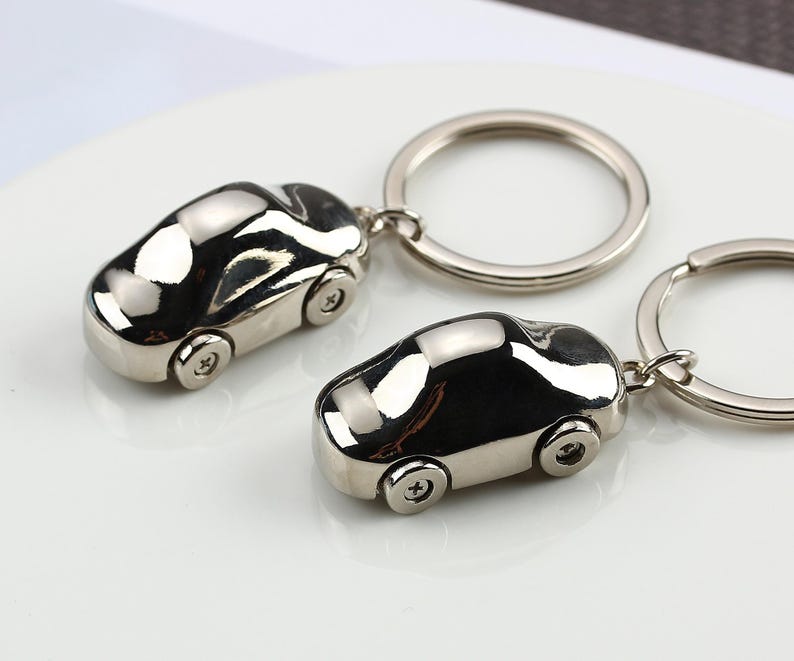 K&ouml;nnte beinhalten: Zwei silberne Autoschl&uuml;sselanh&auml;nger. Jeder Schl&uuml;sselanh&auml;nger zeigt ein Miniaturauto mit detaillierten R&auml;dern und einer glatten, reflektierenden Oberfl&auml;che. Die Schl&uuml;sselanh&auml;nger sind an silbernen Ringen befestigt.