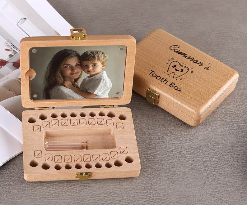 May include: Wooden tooth box with a hinged lid, featuring a photo of a mother and child. The open box reveals spaces for baby teeth and a small vial. The closed lid is engraved with "Cameron's Tooth Box".