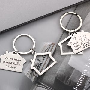 May include: Silver house-shaped keyrings. One keyring reads "Our first Home Becca & Robert 3.24.2024". Another keyring reads "Our New Home" with a drawing of a family, sun, and birds. The keyrings have a circular ring.
