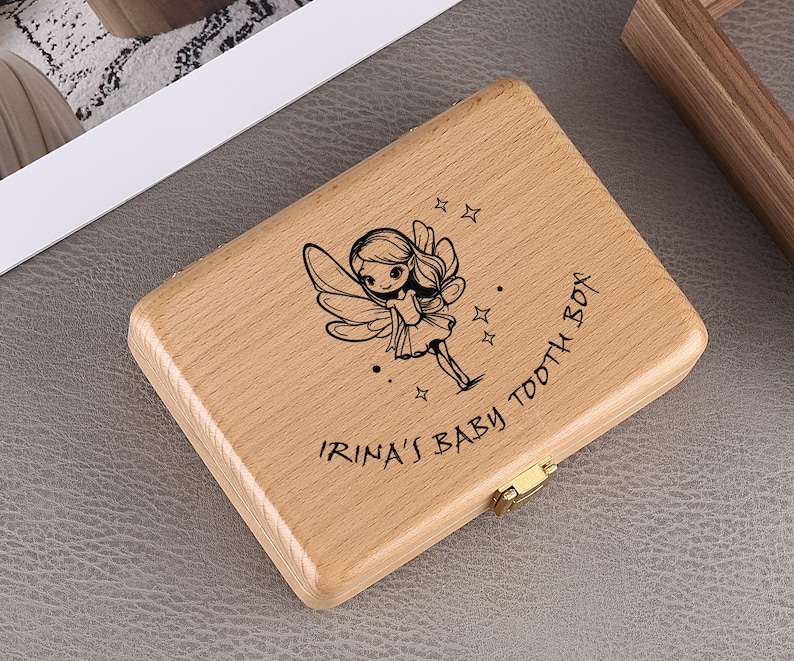 May include: A light-colored wooden baby tooth box with a hinged lid and gold-colored clasp. The box is engraved with a fairy illustration and the text "IRINA'S BABY TOOTH BOX".