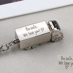 Könnte beinhalten: Ein silberfarbener, truckförmiger Schlüsselanhänger mit der Gravur "Be safe, We love you!" und einem Herzsymbol. Der Schlüsselanhänger hat einen Kettenanhang.