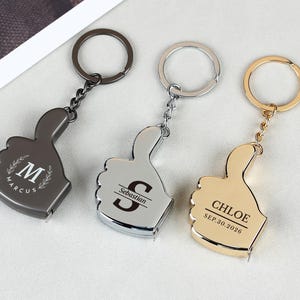 以下が含まれることがあります： 親指を立てた形の金属製キーホルダーメジャー3点。ガンメタル、シルバー、ゴールドの仕上げ。ガンメタルには「M」と「Marcus」、シルバーには「S」と「Sebastian」、ゴールドには「CHLOE」と「SEP.30.2026」の文字が刻印されています。
