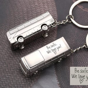 May include: Two silver-colored bus keychains. Each keychain features a detailed bus design with wheels and windows. One keychain has the text "Be safe, We love you!" engraved on it. The keychains are attached to silver key rings.