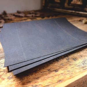 May include: Three dark grey sheets of paper with curved edges, stacked on a rustic wooden surface. The paper has a textured finish. The background includes wooden elements and a decorative item.