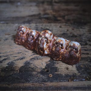 May include: A brown ceramic sculpture featuring a series of five stylized faces with open eye sockets. The sculpture has a textured, organic appearance and is set against a rustic wooden background.