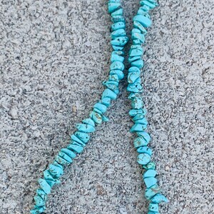May include: A turquoise chip bead necklace with black onyx accents and a skull pendant. The necklace features a toggle clasp and is displayed on a textured gray surface. The turquoise beads are a vibrant blue-green color.