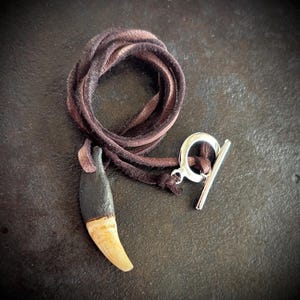 May include: A brown leather cord necklace with a silver toggle clasp and a pendant shaped like a tooth with a black resin coating.