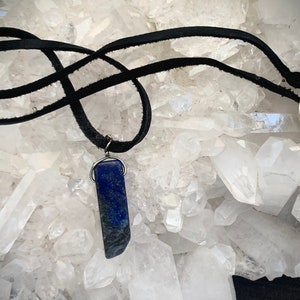 May include: A handcrafted necklace featuring a rectangular, deep blue lapis lazuli pendant. The pendant is suspended from a black suede cord with a silver-tone metal clasp. The necklace is displayed against a backdrop of clear quartz crystals.