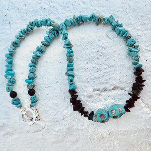 May include: A turquoise stone necklace with black stone accents and two turquoise skull beads. The necklace has a silver toggle clasp. The stones are arranged in a beaded pattern.