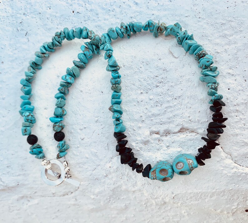 May include: A turquoise stone necklace with black stone accents and two turquoise skull beads. The necklace has a silver toggle clasp. The stones are arranged in a beaded pattern.