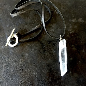 May include: A long, clear quartz crystal pendant hangs from a black leather cord. The cord has a silver toggle clasp.