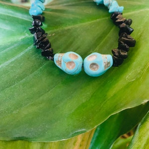 May include: A beaded bracelet featuring turquoise skull beads and black stone chips. The bracelet is strung on a black cord and is displayed on a green leaf.