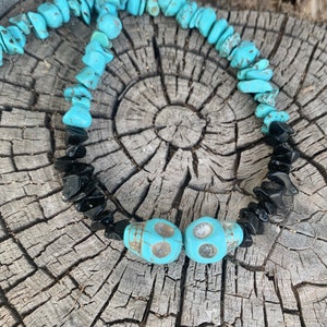 May include: A turquoise and black beaded necklace with two turquoise skull beads. The necklace features small, irregular turquoise and black beads. The necklace is displayed on a weathered, gray wooden surface.