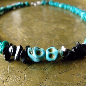 May include: A turquoise and black beaded necklace with two turquoise skull beads. The necklace features small, irregular turquoise beads and black stone chips. The necklace is secured with a silver-colored clasp.