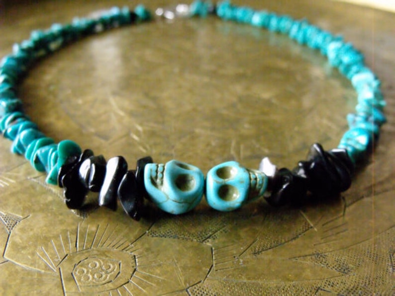 May include: A turquoise and black beaded necklace with two turquoise skull beads. The necklace features small, irregular turquoise beads and black stone chips. The necklace is secured with a silver-colored clasp.