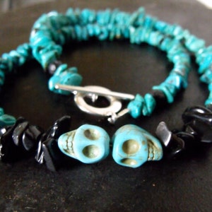 May include: A turquoise stone necklace with black stone accents and two turquoise skull beads. The necklace has a silver-colored toggle clasp. The turquoise stones are irregularly shaped.