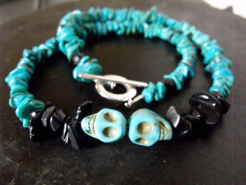 May include: A turquoise stone necklace with black stone accents and two turquoise skull beads. The necklace has a silver-colored toggle clasp. The turquoise stones are irregularly shaped.