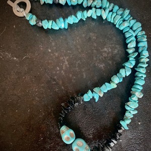 May include: A turquoise stone necklace with a silver toggle clasp. The necklace features turquoise chip beads, black stone chips, and two turquoise skull beads. The necklace is displayed on a dark surface.
