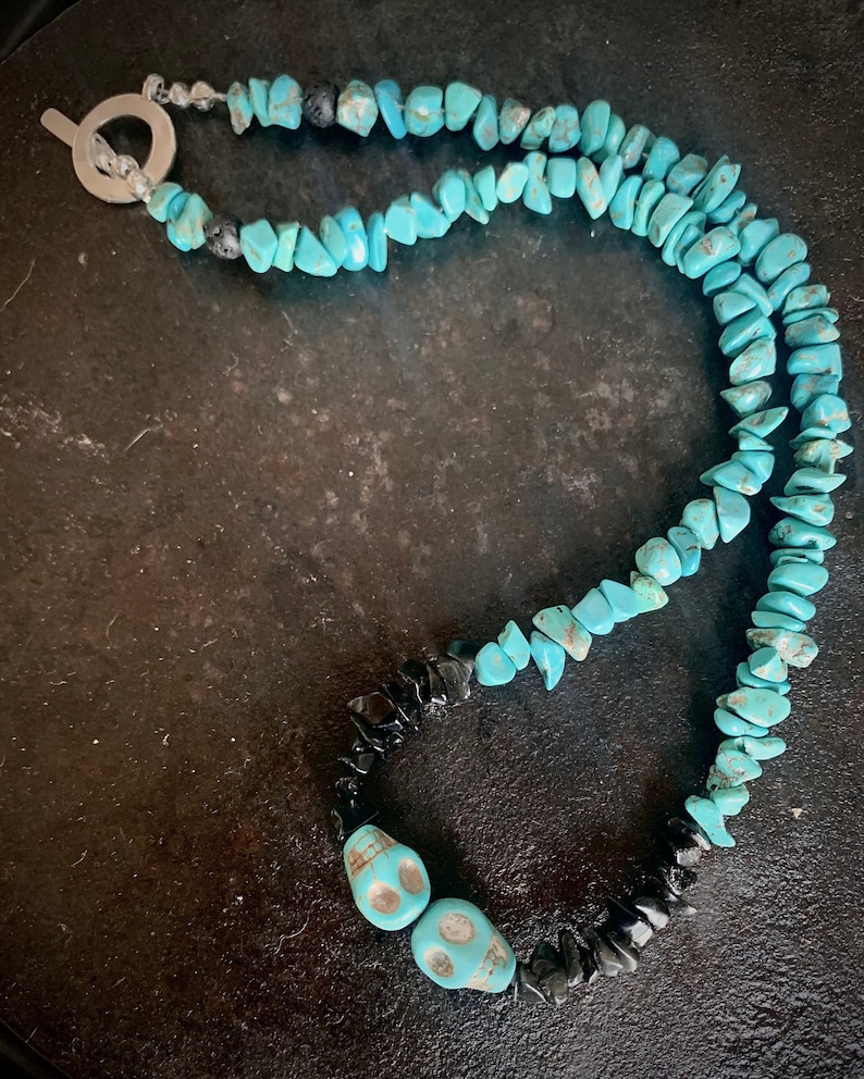 May include: A turquoise stone necklace with a silver toggle clasp. The necklace features turquoise chip beads, black stone chips, and two turquoise skull beads. The necklace is displayed on a dark surface.