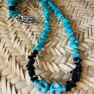 May include: A turquoise and black beaded necklace with two turquoise skull beads. The necklace features a silver clasp and is displayed on a woven straw surface. The necklace is a unique piece of jewelry.