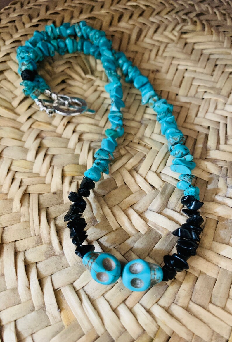 May include: A turquoise and black beaded necklace with two turquoise skull beads. The necklace features a silver clasp and is displayed on a woven straw surface. The necklace is a unique piece of jewelry.