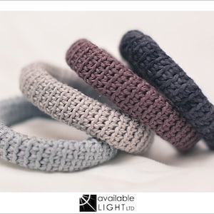 Pode incluir: Quatro pulseiras de malha em tons de cinza, malva e azul marinho são dispostas sobre uma superfície branca. As pulseiras têm uma aparência espessa e texturizada, com um padrão de malha visível. As pulseiras são empilhadas e ligeiramente anguladas.