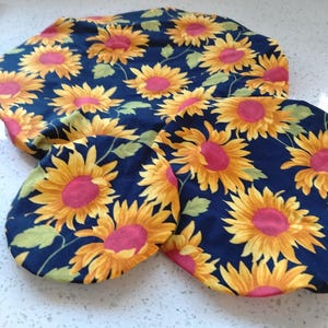 May include: A set of kitchen accessories with a sunflower design. The items include a round pot holder, a square pot holder, and oven mitts. The sunflowers have yellow petals and pink centres on a dark blue background.