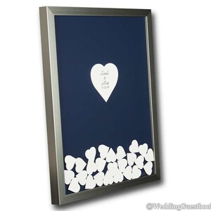 May include: A silver framed wedding guest book with a navy blue background. The book features a large white heart with the names "Sarah & Matt" and the date "3-12-18" written on it. The bottom of the frame is filled with smaller white hearts for guests to sign.