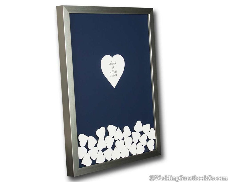 May include: A silver framed wedding guest book with a navy blue background. The book features a large white heart with the names "Sarah & Matt" and the date "3-12-18" written on it. The bottom of the frame is filled with smaller white hearts for guests to sign.