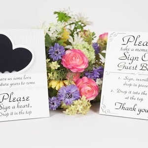 May include: Two white signs with black text and decorative borders. The sign on the left has a black heart shape with a white lace border. The text on the left sign reads: "Leave us some love for future years to come. Please sign a heart, drop it in the top." The sign on the right reads: "Please take a moment to sign our guest book. 1. Sign, inscribe a drop-in piece. 2. Drop it into the frame at the top. Thank you!"