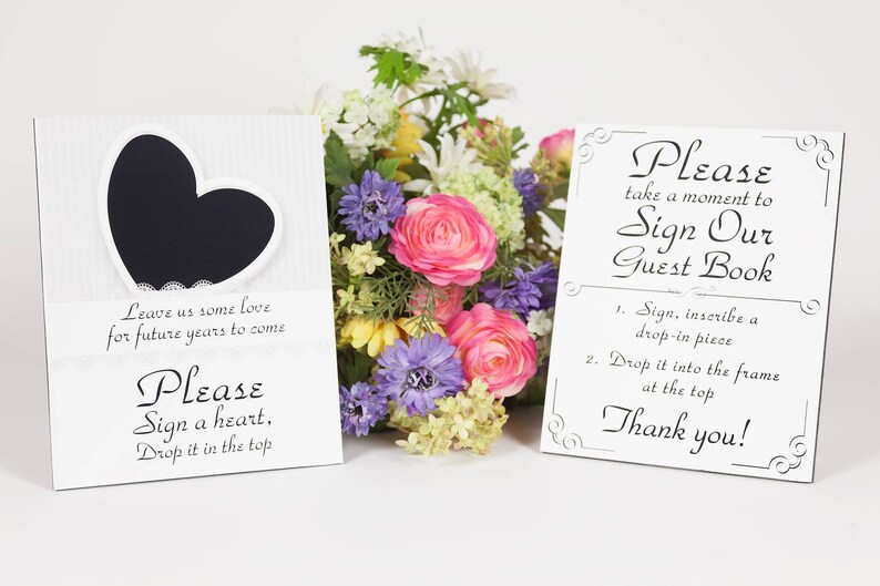 May include: Two white signs with black text and decorative borders. The sign on the left has a black heart shape with a white lace border. The text on the left sign reads: "Leave us some love for future years to come. Please sign a heart, drop it in the top." The sign on the right reads: "Please take a moment to sign our guest book. 1. Sign, inscribe a drop-in piece. 2. Drop it into the frame at the top. Thank you!"