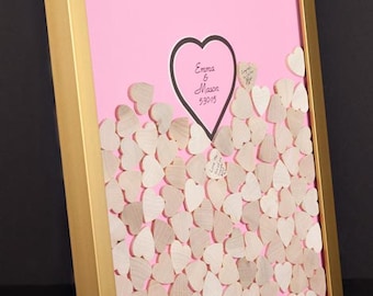 Peut inclure: Une image encadrée en or avec un fond rose. L'image présente une découpe en forme de cœur avec le texte "Emma & Mason 5.30.15" à l'intérieur. Le fond est rempli de petits cœurs en bois.