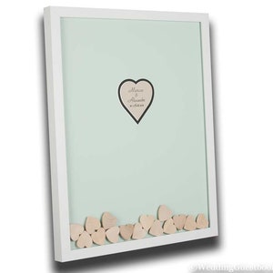 May include: White frame with a light green background. A heart-shaped cutout with the text "Marion & Alexandra 10.07.2019" is in the center. The frame is filled with small wooden hearts.