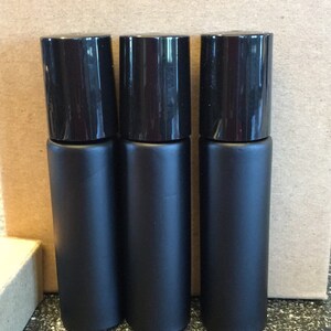 May include: Three black glass roller bottles with black caps. The bottles are standing on a dark speckled surface.