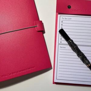 May include: A pink leather journal with the text "The Little Big Journal Company" embossed on the cover. A separate pink notepad with a to-do list and notes section is open and a black pen with a silver dragon design is lying on top of it.