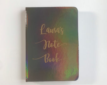 Op de afbeelding: Een zwart, iriserend notitieboek met gouden letters die "Laura's Note Book" lezen.