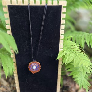 Puede incluir: Un collar con colgante artesanal con un colgante redondo de madera con un diseño azul y blanco. El colgante cuelga de un cordón negro. El collar se exhibe sobre una superficie de terciopelo negro, con un expositor de madera y follaje verde.